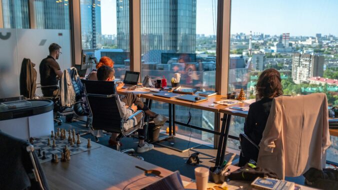 Et lyst og moderne kontormiljø med store vinduer, flere medarbejdere, der arbejder ved skriveborde, og en imponerende udsigt over byen i baggrunden.